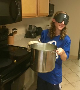 a young woman wearing sleep shades carries a large pot of boiling pasta to the strainer at the sink