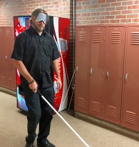 A man with a white cane finds his way past lockers and soda machine wearing sleepshades