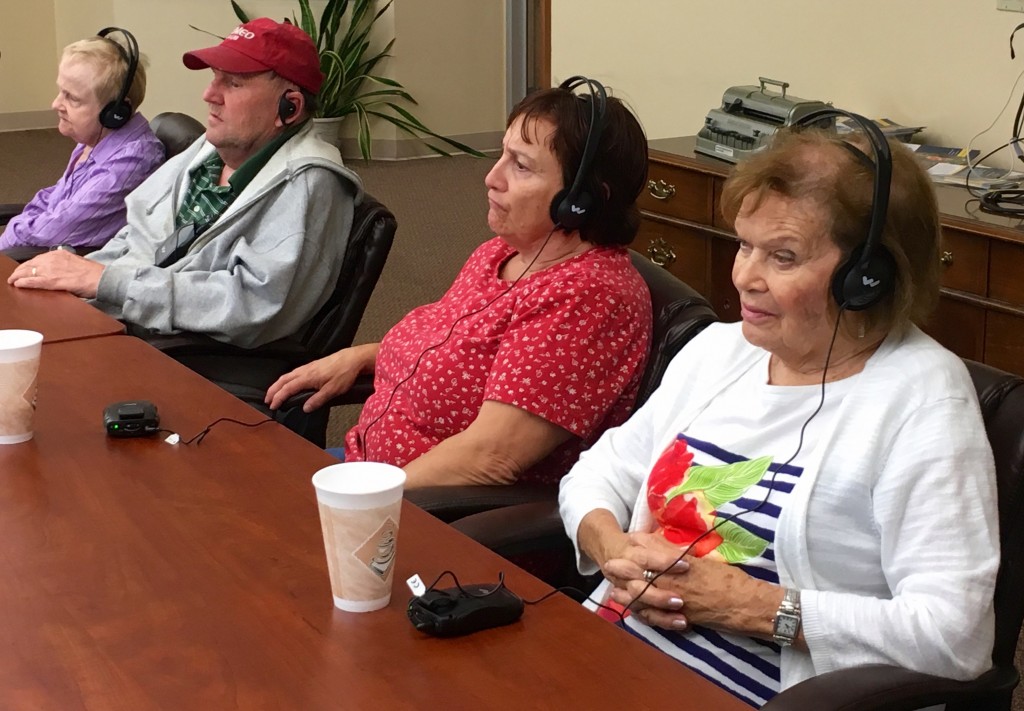 Four Seniors with earphones listen intently, pocket-sized receivers on the table in front of them.