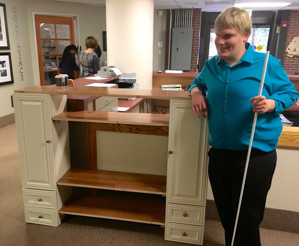 a grinning young man leans an elbow on top of the entertainment center he built, white cane in his other hand