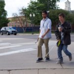 a young man and a teenager with cane and sleepshades cross a suburban intersection