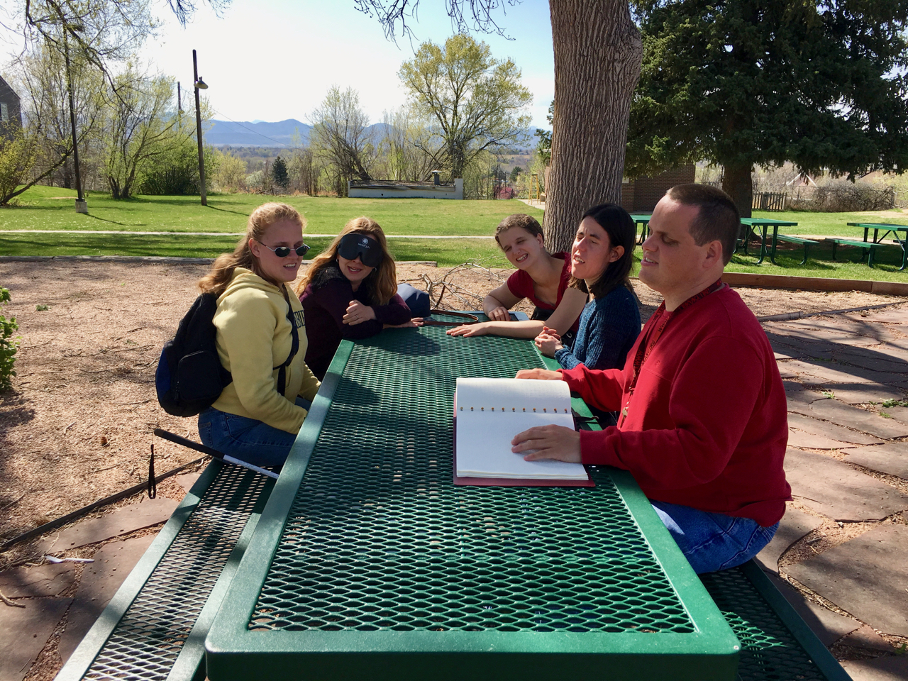 Five people around a picnic table, one reading Braille aloud to the others.