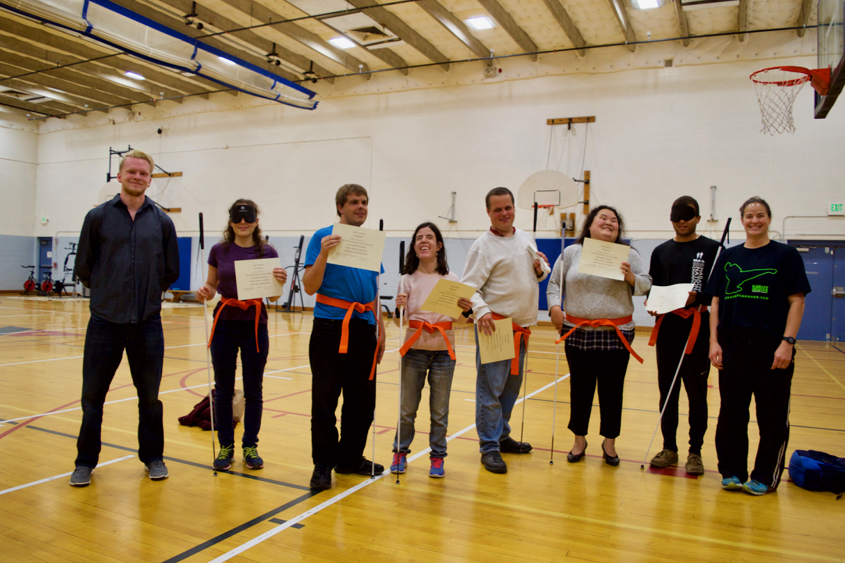 six class members stand in a semi-circle wearing their new Orange belts and hold certificates, flanked by two Karate Denver instructors