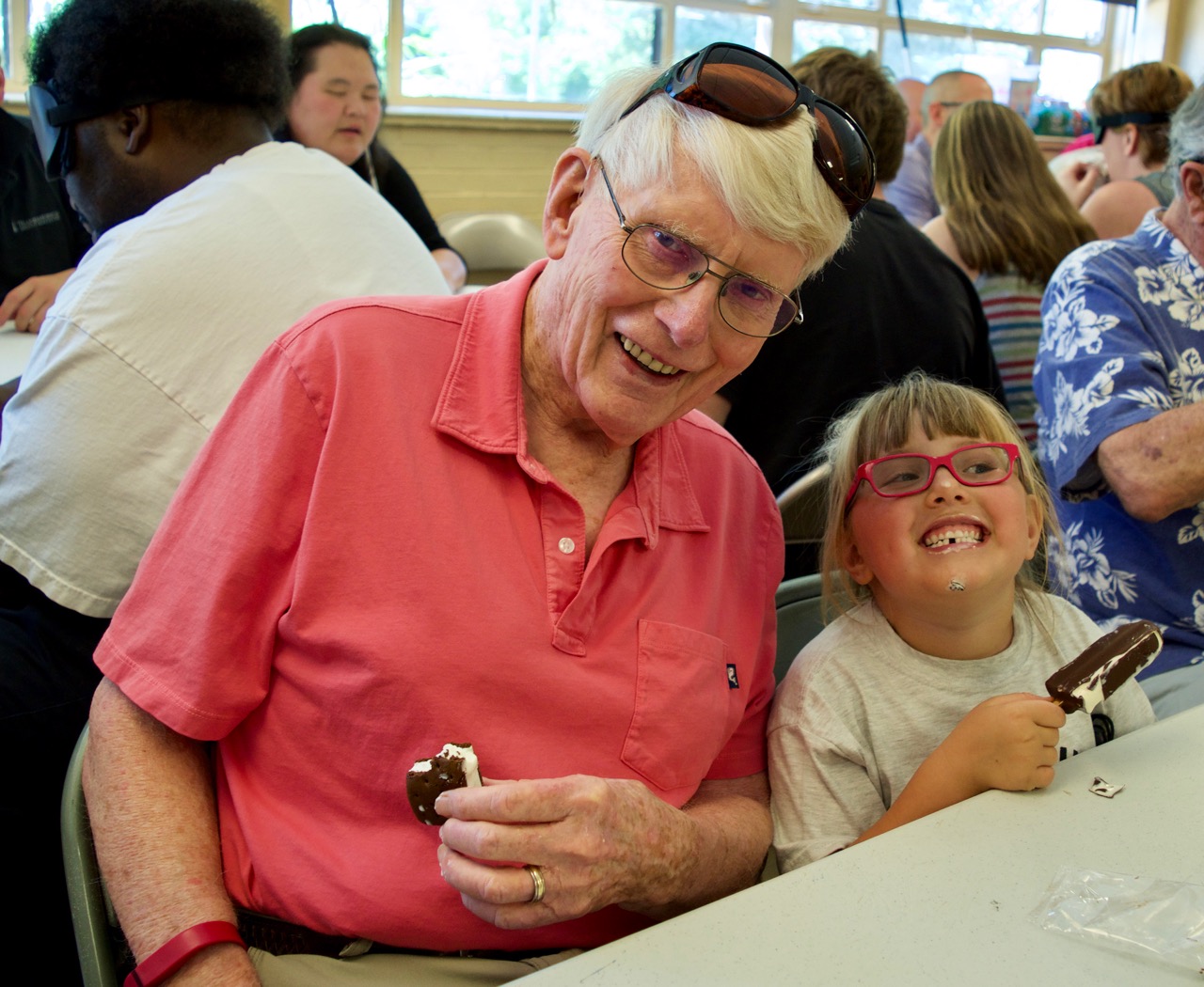 A man in his eighties sits beside a 6-year-old girl, both enjoying ice cream