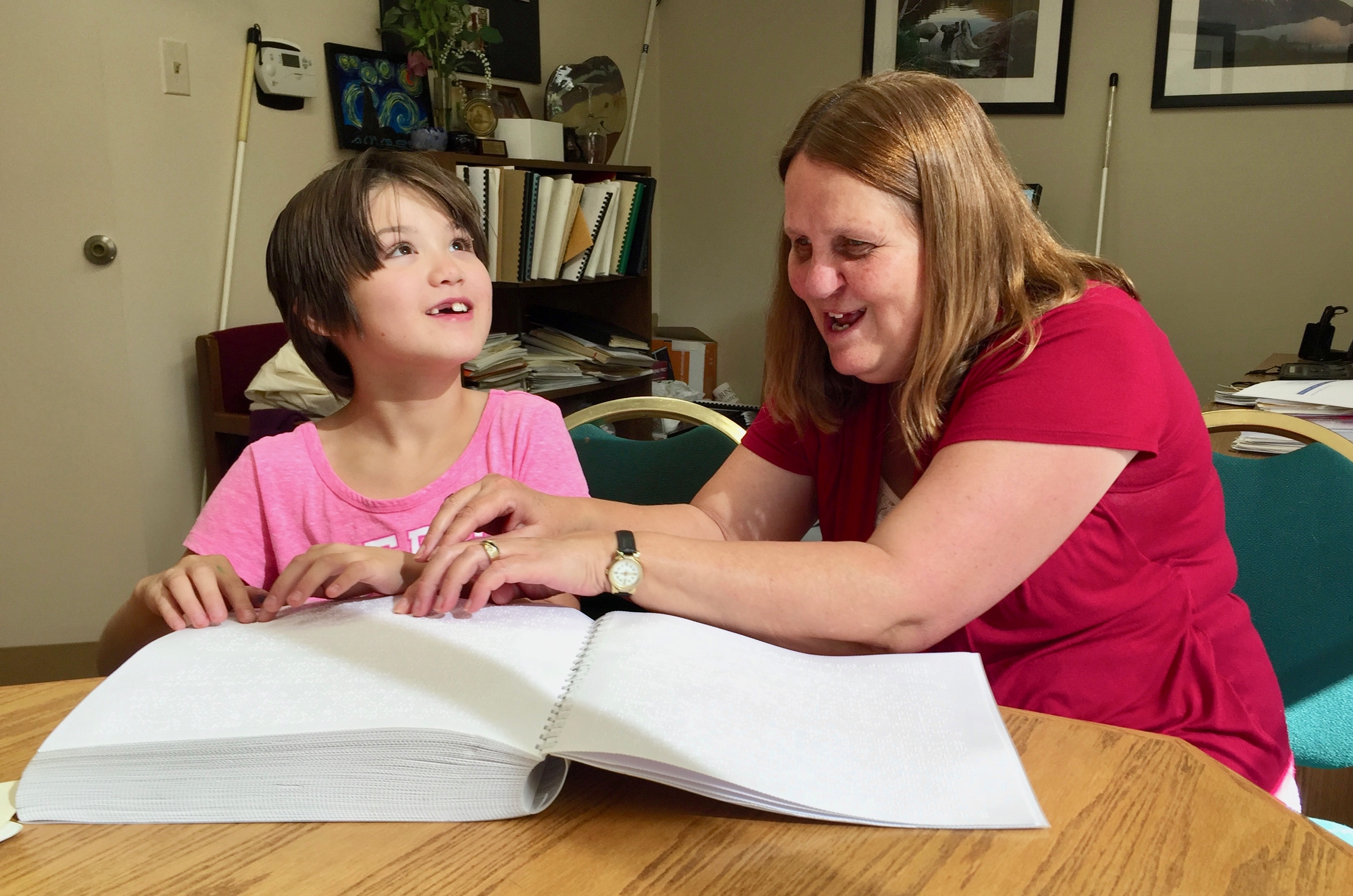a smiling girl with missing front teeth and a woman with their hands on the same page of Braille