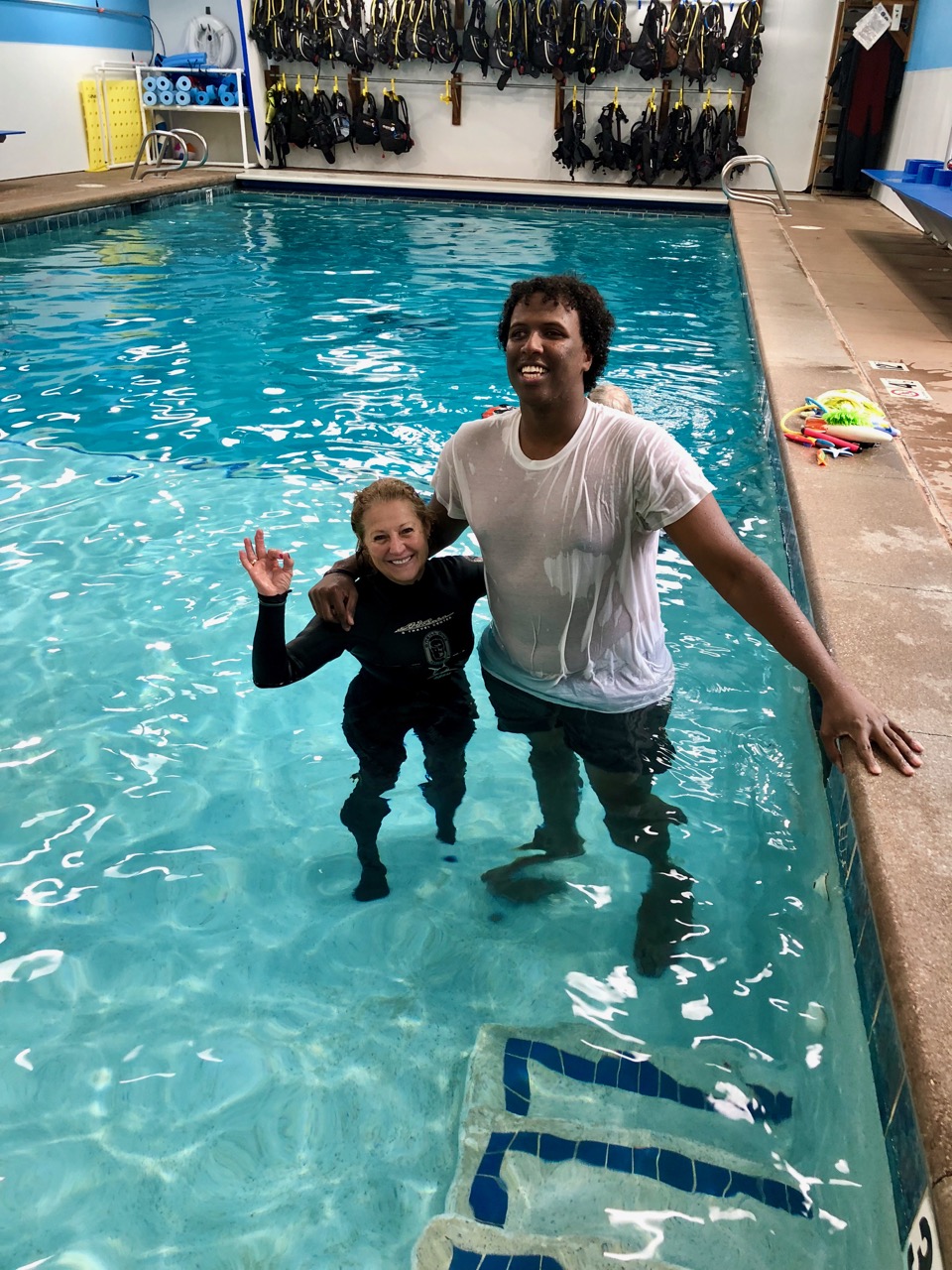 A tall young man in t-shirt stands with his petite, wet-suited instructor in the shallow end of the pool