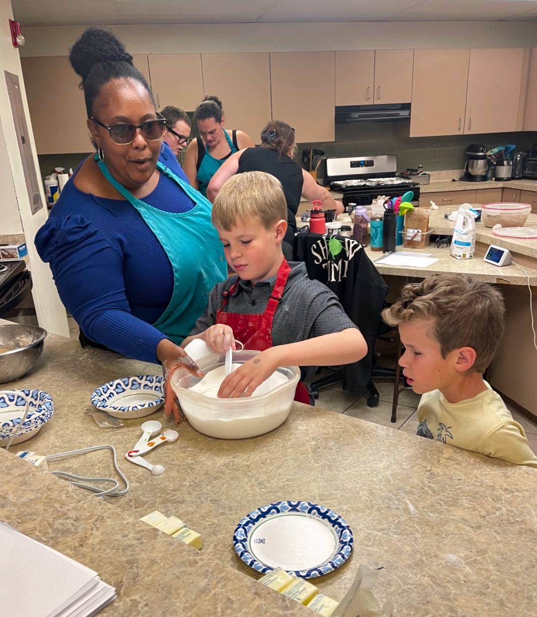 A young woman with her hair in a bun guides two school-aged boys in mixing cookie dough in a busy kitchen.