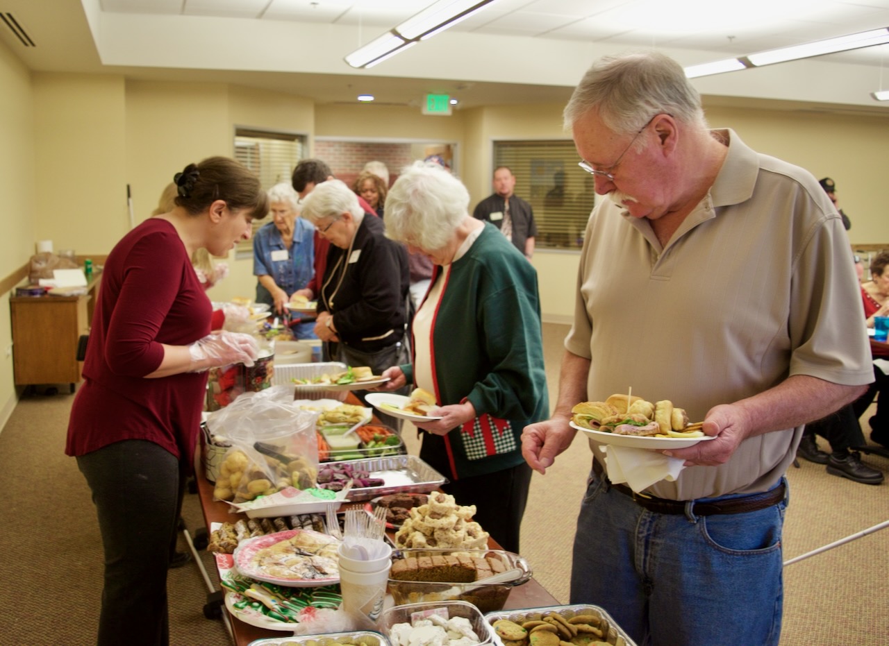 A woman reaches across a long table filled with food to place something on an older man’s plate.