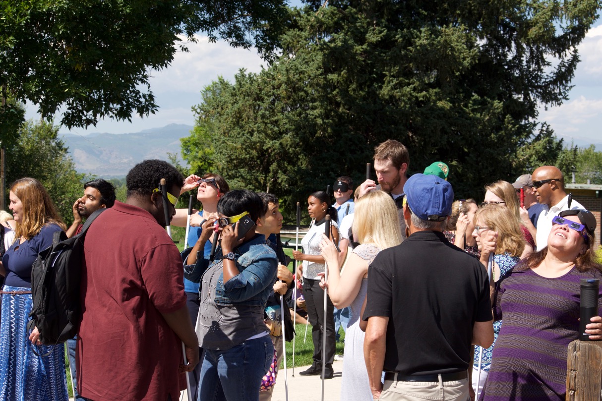 A large group of CCB staff and students outside observing the eclipse. Some are wearing eclipse glasses while they listen to a live audio description