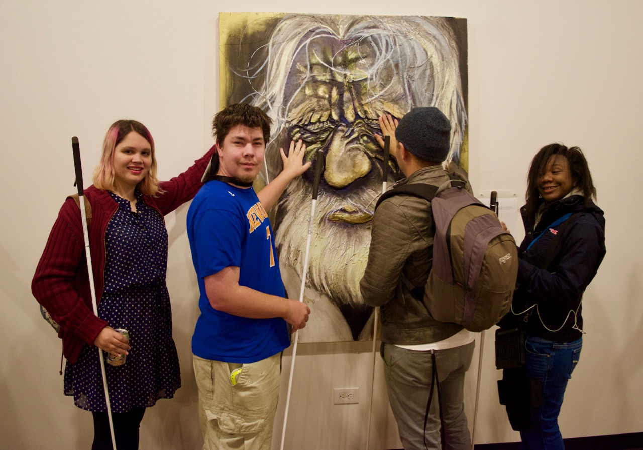 Adia, Mason M. and Cezar look at the Old Man Wincing while Ravi reads the description provided in Braille