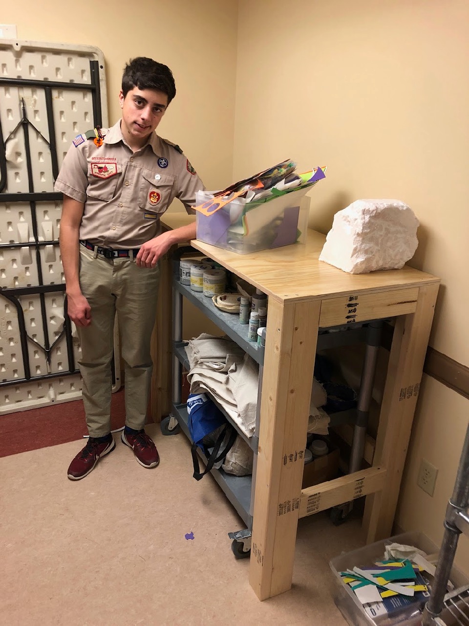 Alex LaBarre stands next to one of the newly completed work benches showing how a rolling art supply cart fits underneath