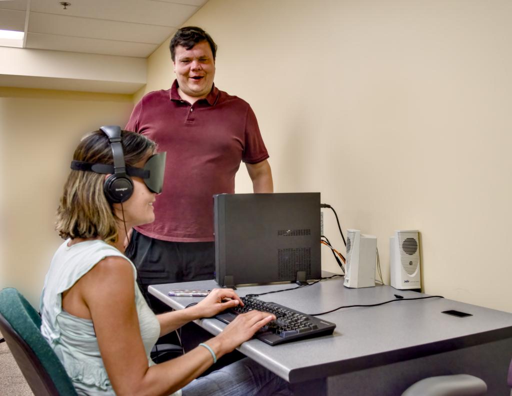 A woman in a classroom sits at a desk with a computer while a man stands behind her, offering support.
