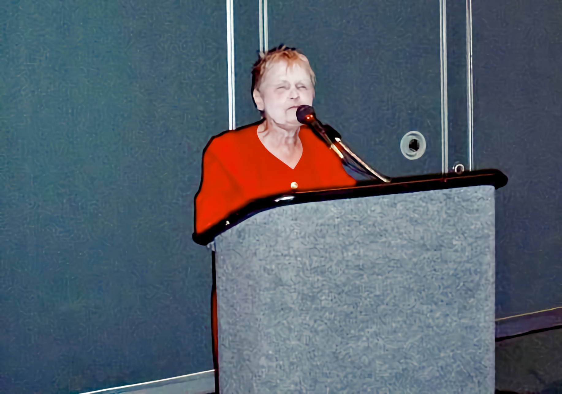 a small well-dressed woman in her 70s stands at a podium, microphone near her face