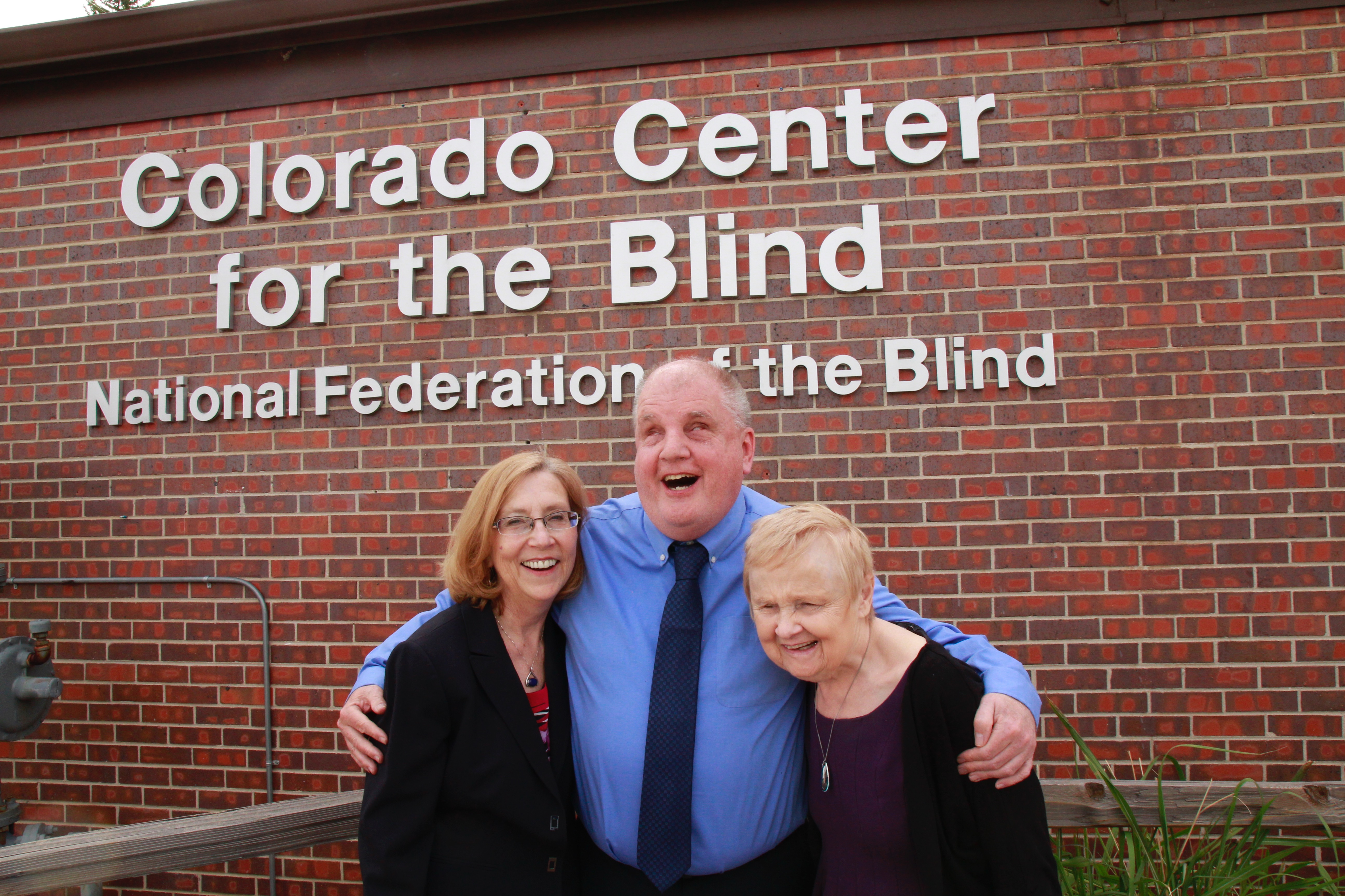Duncan, Tom and Diane hug in front of the CCB sign on a bright spring