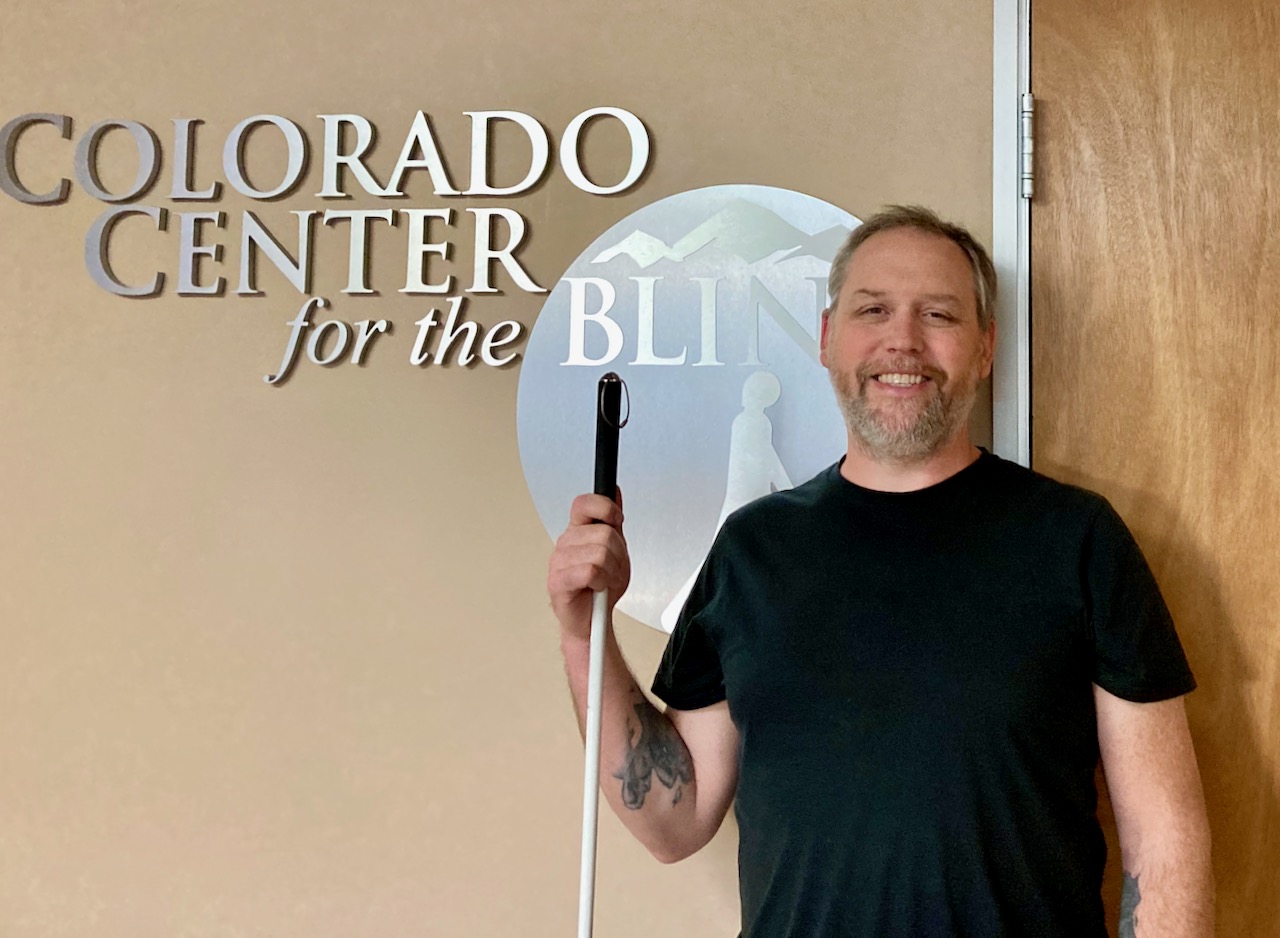 Fitz stands and smiles with his White Cane near the CCB Logo