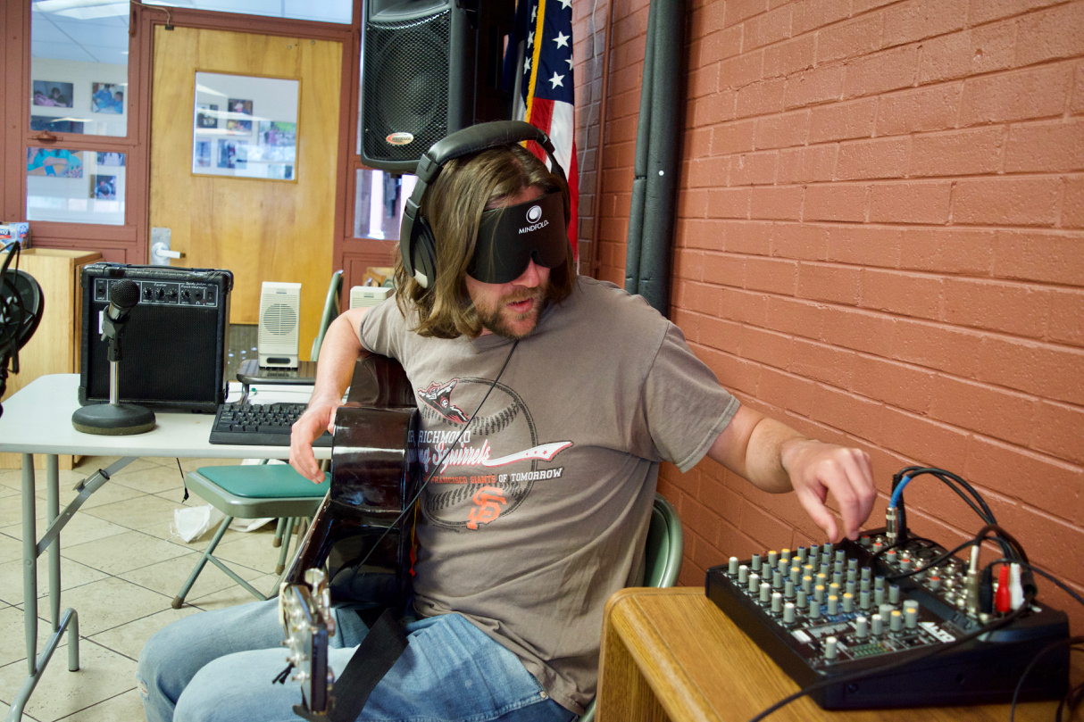 Graham’s hands turn dials on the audio mixing board, mic, headphones and guitar visible