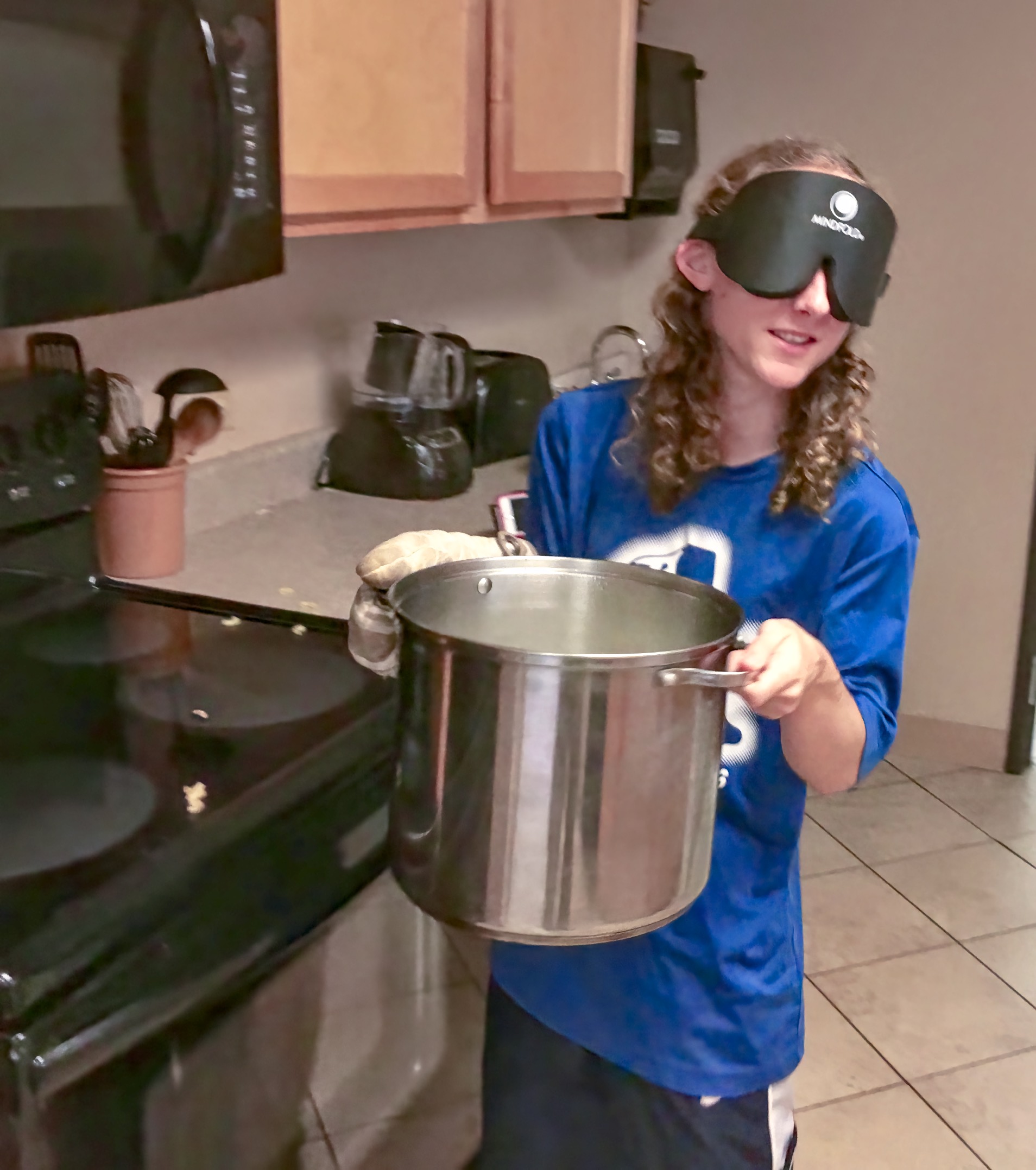 a young woman wearing sleep shades carries a large pot of boiling pasta to the strainer at the sink