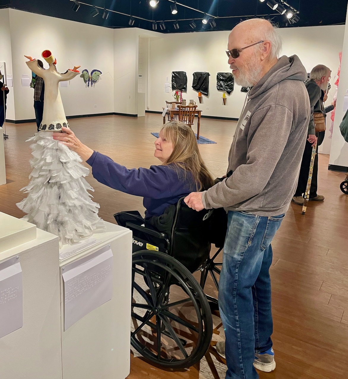 A man holds the hand grips of his wife’s wheelchair as she reaches up to touch a ceramic piece