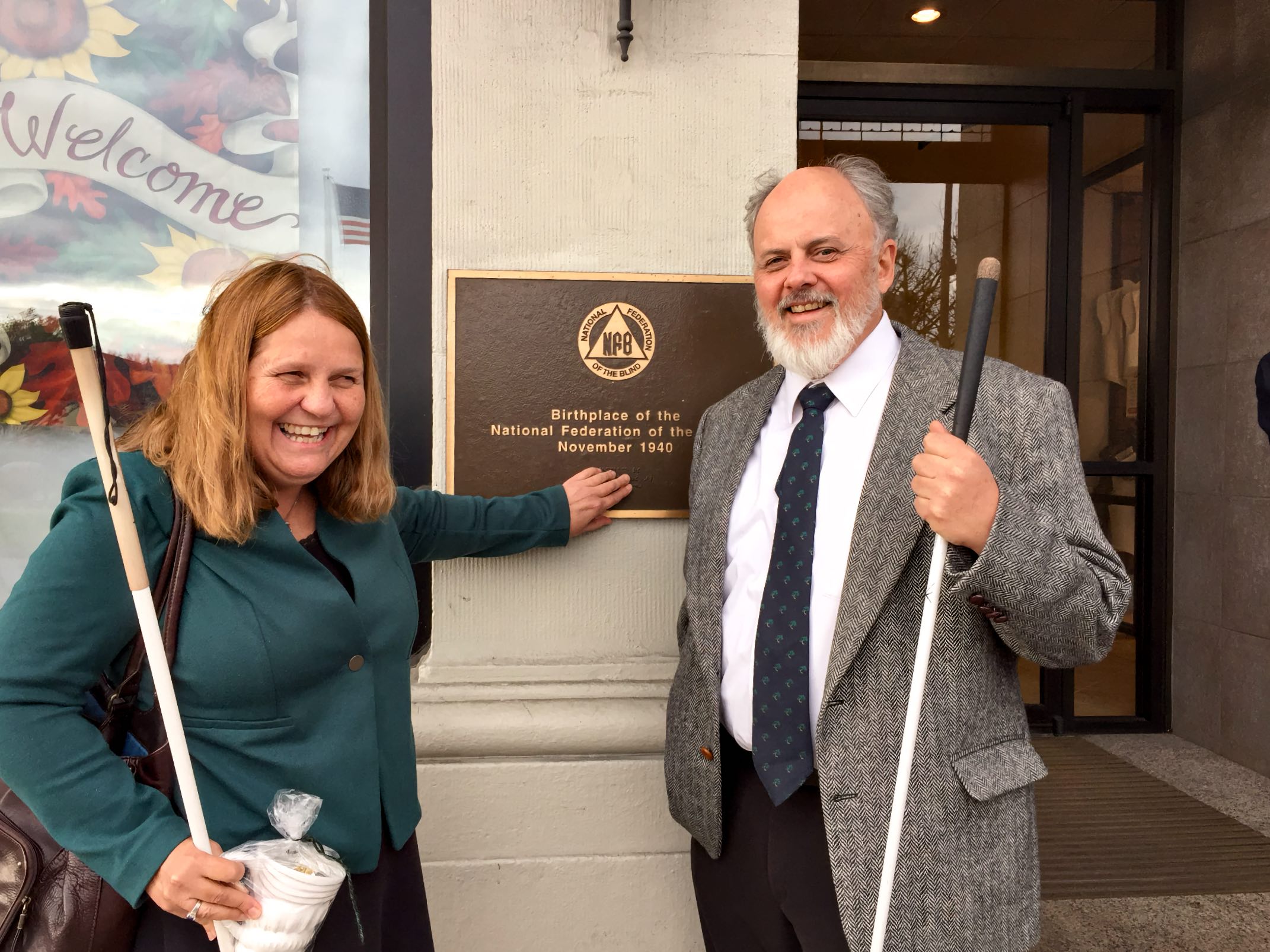 A smiling man and woman stand on each side of a bronze plaque with print and Braille visible. Both hold long white canes.