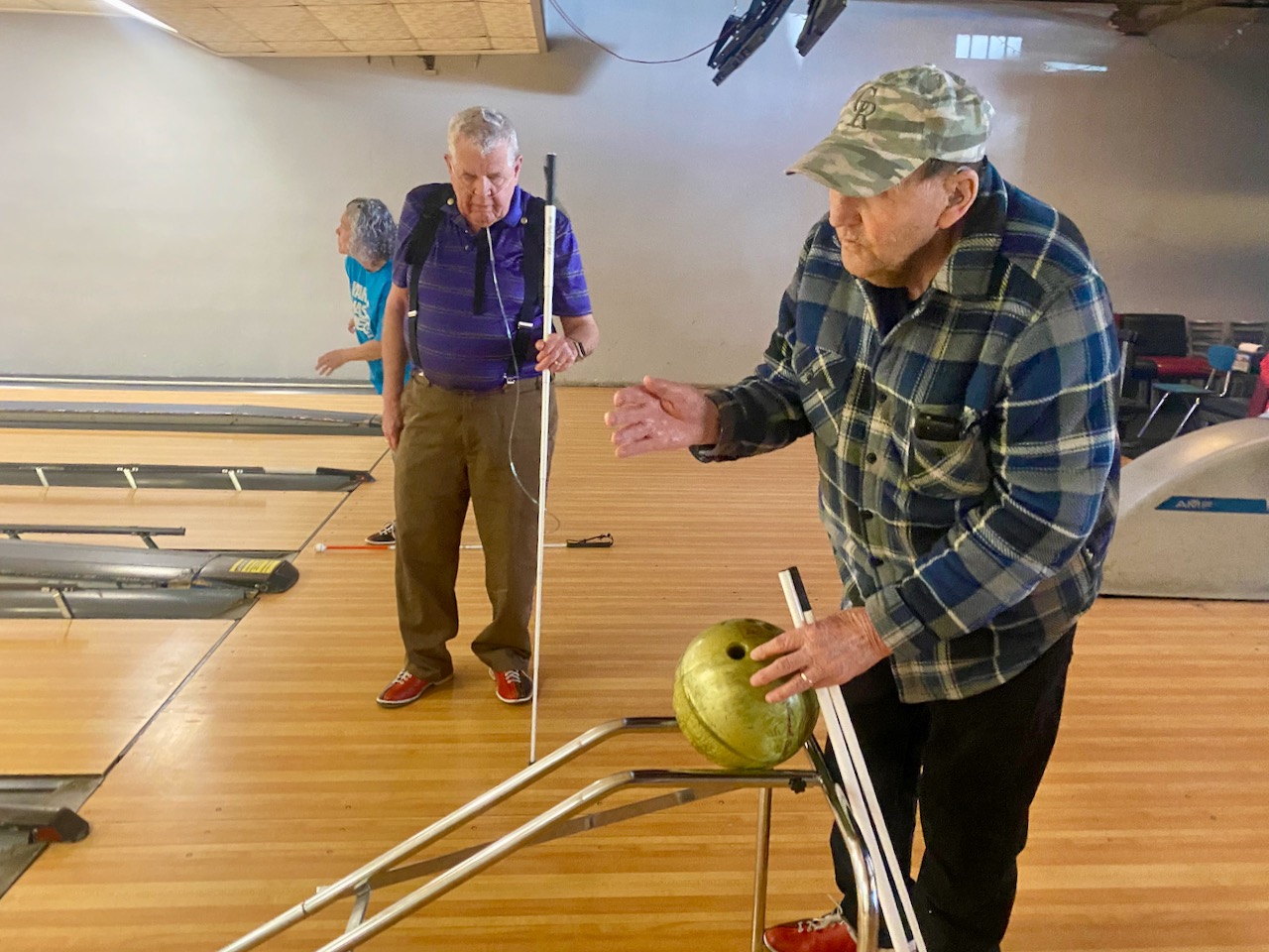 Holding his green ball in place at the top of the angled ramp, John calls his shot as he gestures with his right hand. Dale approaches with his white cane and wearing an oxygen backpack , ready to take his turn.