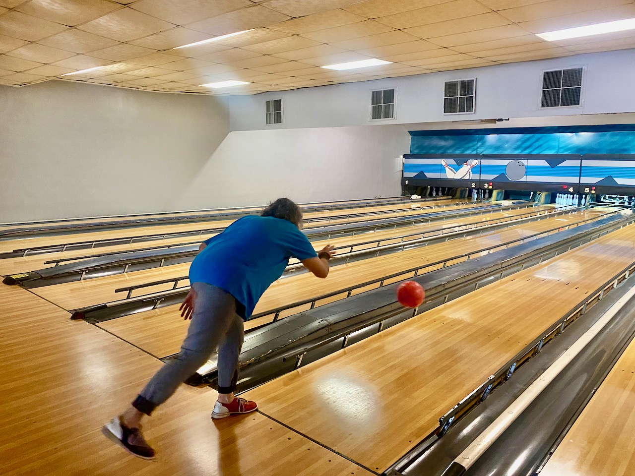 Saray stands with her left foot against the line just after releasing her orange ball, which is still in the air hurtling toward the pins.