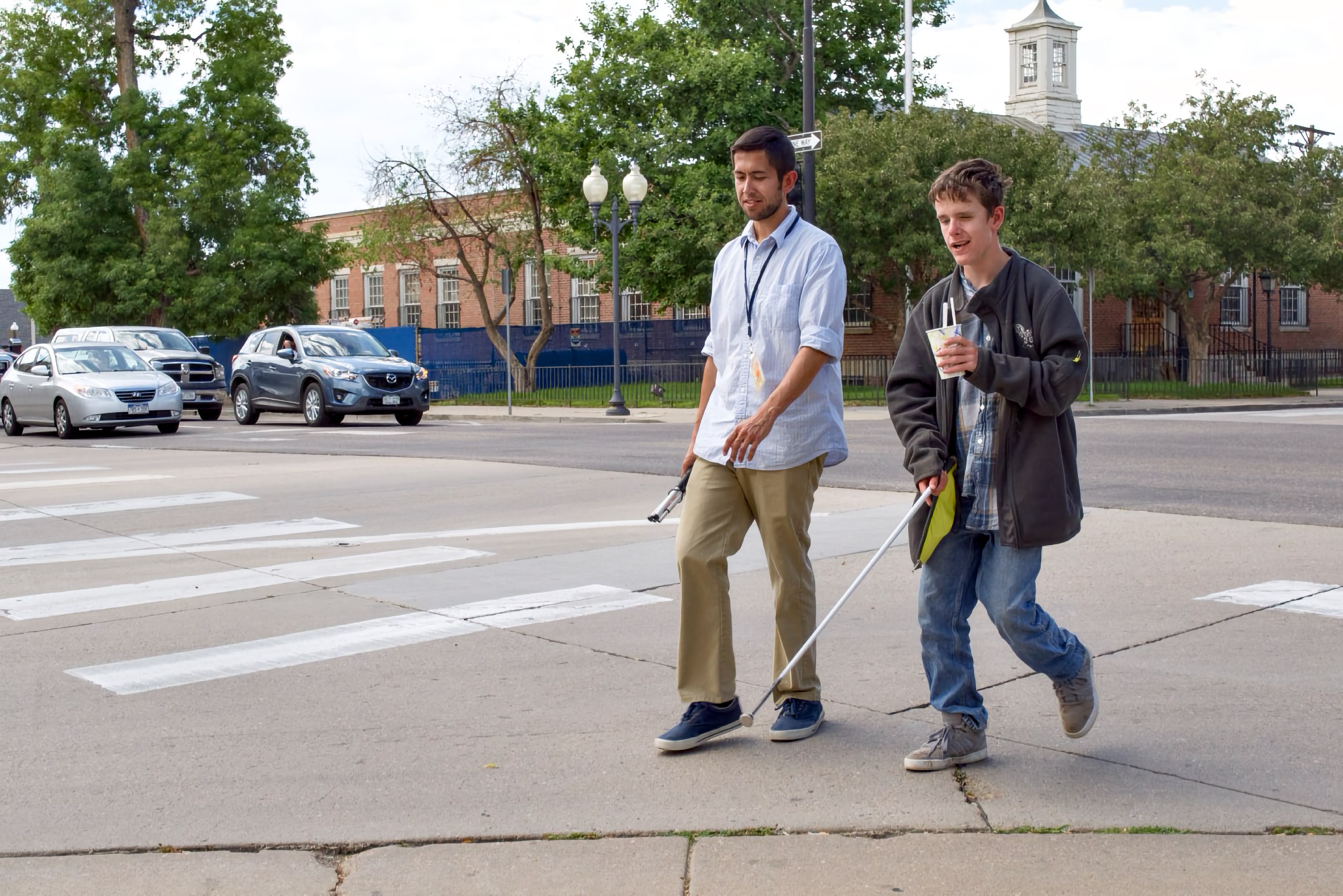 a young man and a teenager with cane and sleepshades cross a suburban intersection