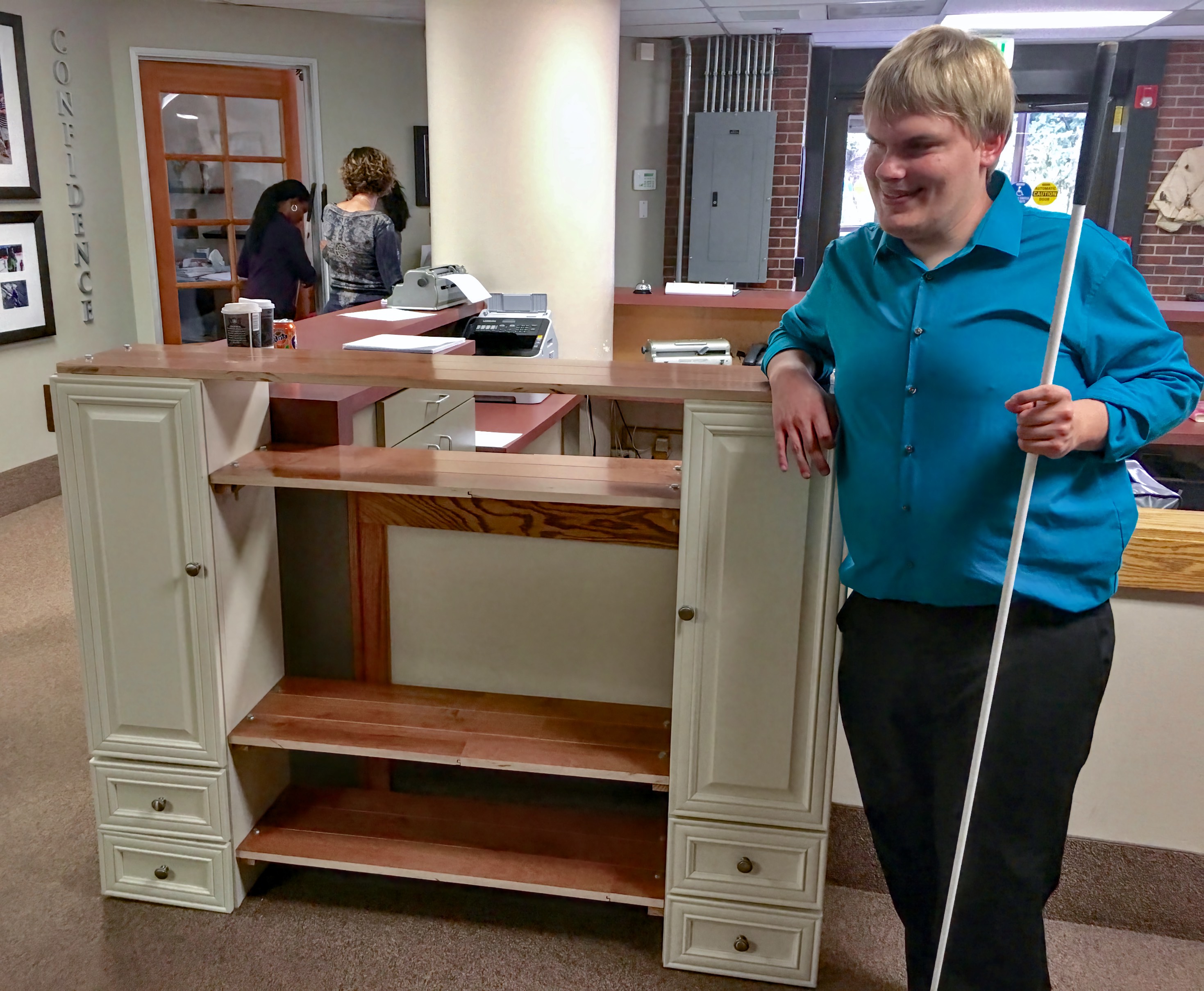 a grinning young man leans an elbow on top of the entertainment center he built, white cane in his other hand