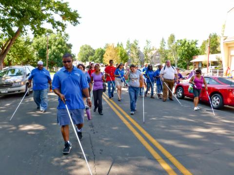 a group of people carrying white canes walk down the middle of a street