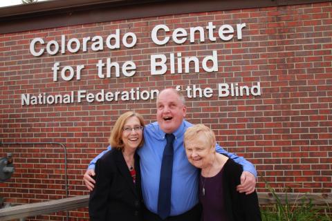 Duncan, Tom and Diane hug in front of the CCB sign on a bright spring
