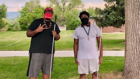Two young Black men wearing COVID masks and holding white canes stand under leafy trees.