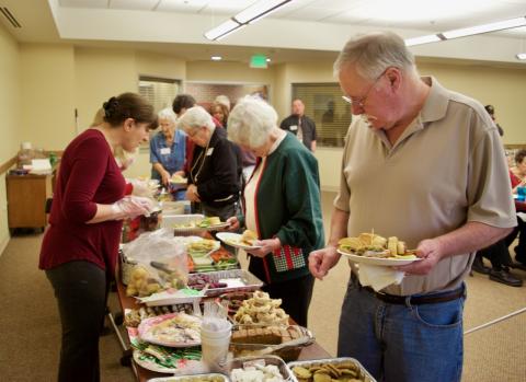 A woman reaches across a long table filled with food to place something on an older man’s plate.