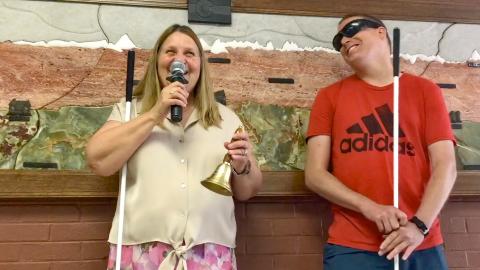 Doug wearing learning shades smiles as he looks at Julie laughing and holding a mic in one hand and a bell in the other.