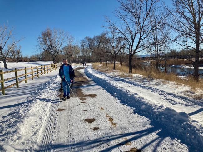 A man in winter clothes and boots extends his cane as he walks along a cleared, but snowy bike-pedestrian path, the South Platte to his right, a pedestrian bridge is further back, and the late-day winter sun on the snowy Rockies in the far distance.