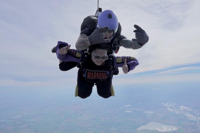 a grinning young woman strapped to her jump-master gives a double thumbs-up while skydiving