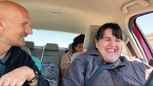 A blind woman is smiling and driving confidently, while her Master Drive instructor, seated next to her, laughs joyfully during an exciting driving lesson.