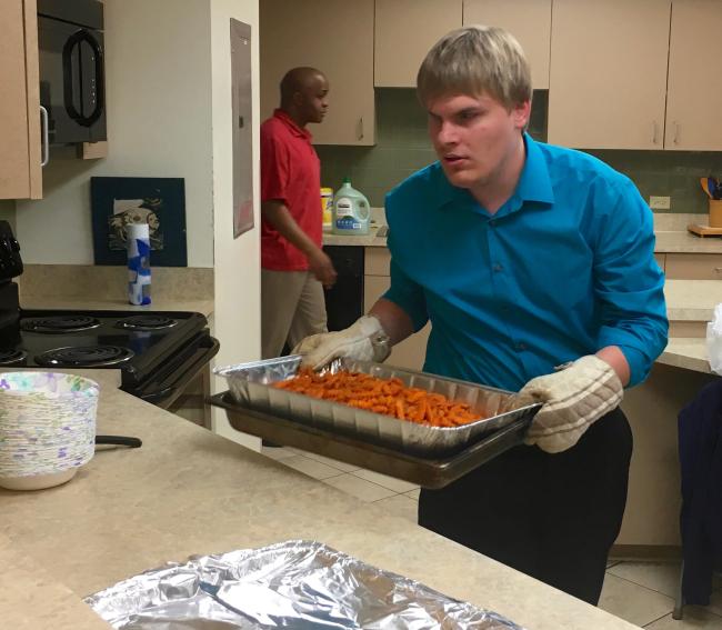 Zach wearing oven mitts carrying a hot tray of french fries into the kitchen about to place it on the serving area