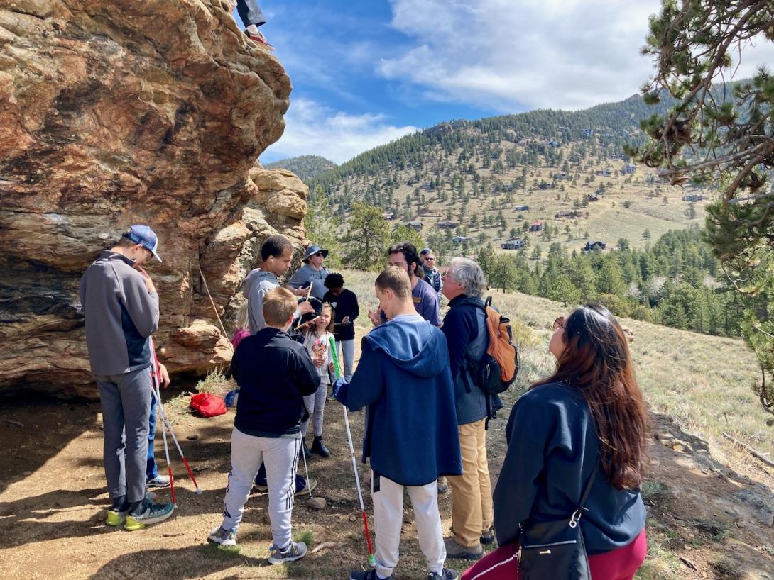 Chaz stands with a group of parents and Blind Youth at the Estes Park Family Weekend 2024, examining an artifact replica near a large rock outcropping.