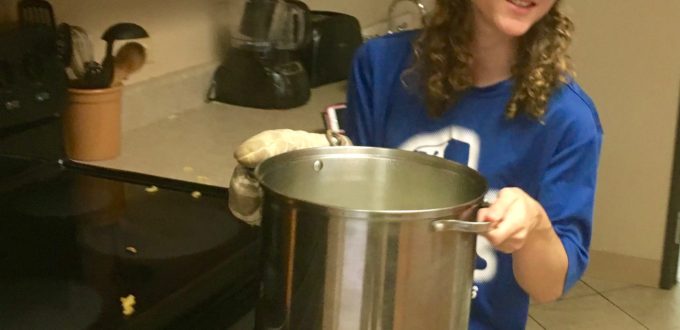 a young woman wearing sleep shades carries a large pot of boiling pasta to the strainer at the sink