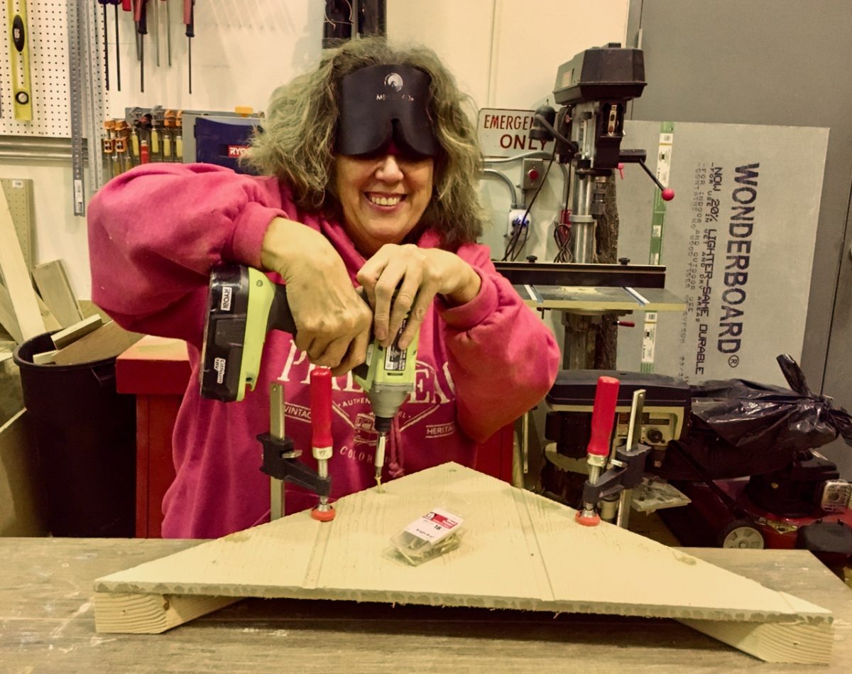 Vicki uses a cordless drill to screw a large trianguler peice onto a frame for her dog house woodshop project