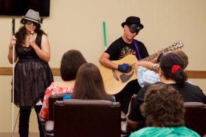 A young woman and young man with guitar perform before a seated crowd. They wear stunning hats.
