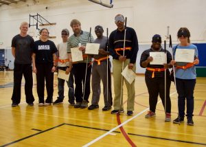 Group shot with instructors standing with the students who are wearing their orange belts and holding their certificates