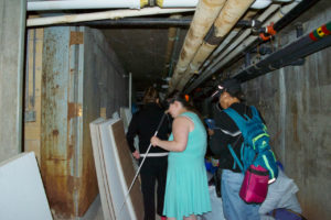A view of a group of students exploring a tight passage way with lots of pipes and supplies stored along the way