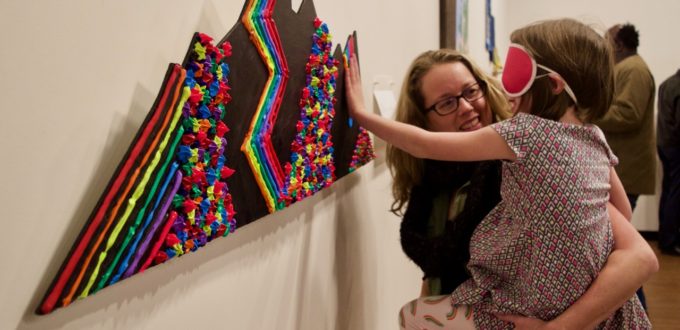 Katy holds up her daughter Zoey who is wearing sleepshades so she can touch ta tactile art piece of the mountains