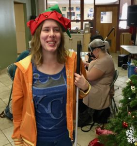 Holly wears a colorful elf hat with bells and smiles while standing next to the Christmas Tree