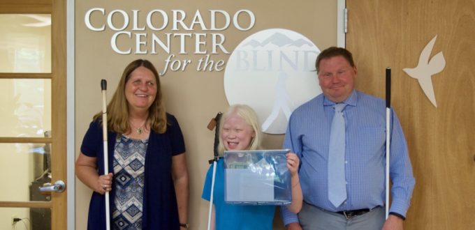 Sixth-grader Chloe stands between Julie and Brent with the tactile CCB logo behind them.