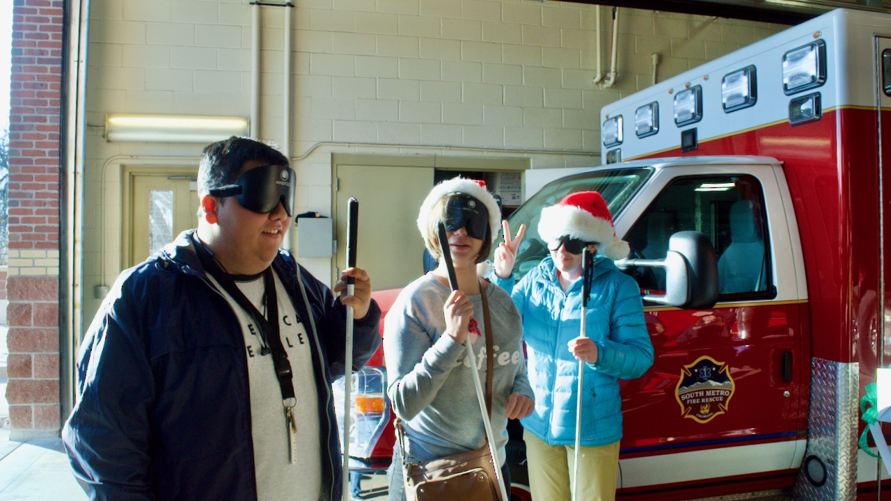 Three young students stand in front of a medical vehicle, all wearing Santa hats