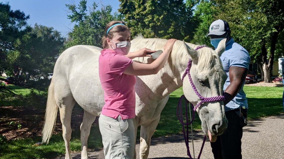 Kelly in sleep shades and mask turns her head to her right while she brushes a white horse named Booger with her left hand. Booger is sticking his tongue out.