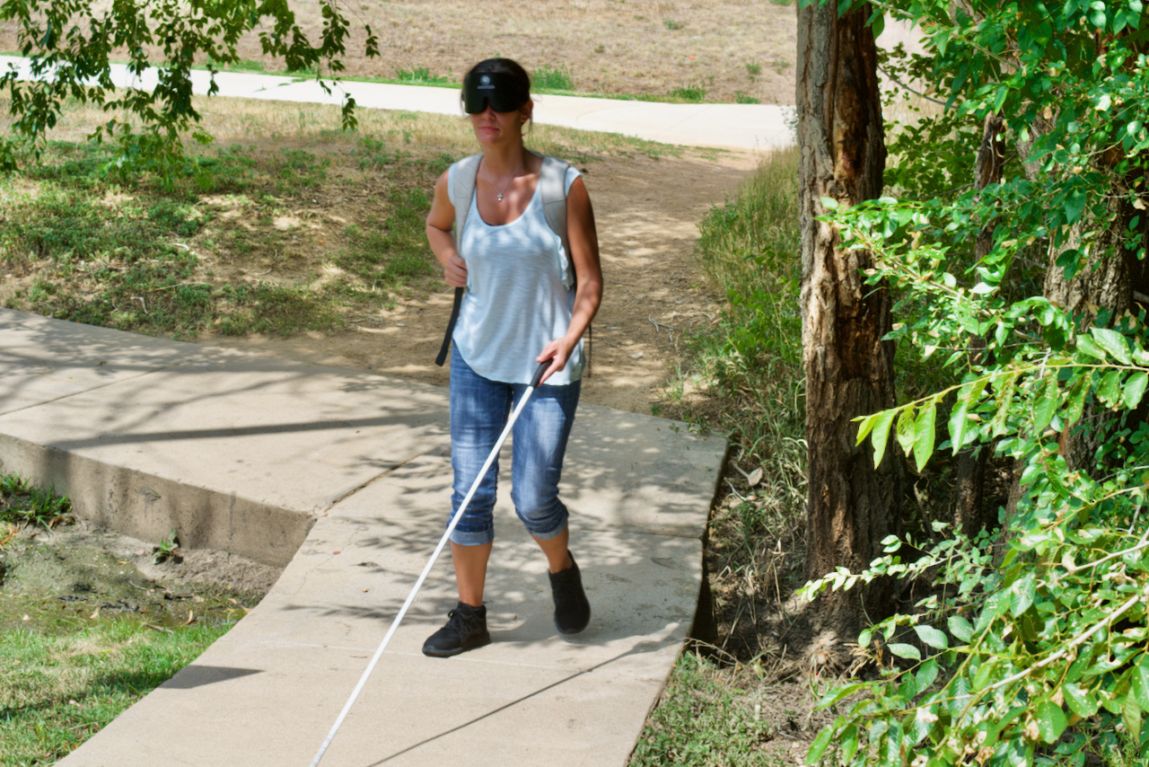 A woman wearing sleepshades and backpack walks uphill on a narrow sidewalk with her white cane