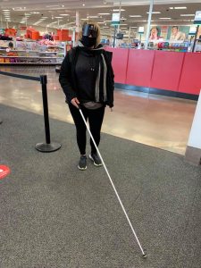 Katie at Target holding her cane and wearing mask and sleep shades during travel class