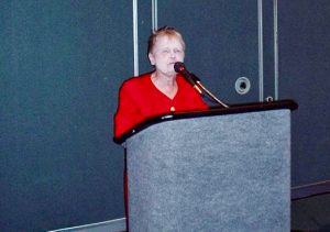 a small well-dressed woman in her 70s stands at a podium, microphone near her face